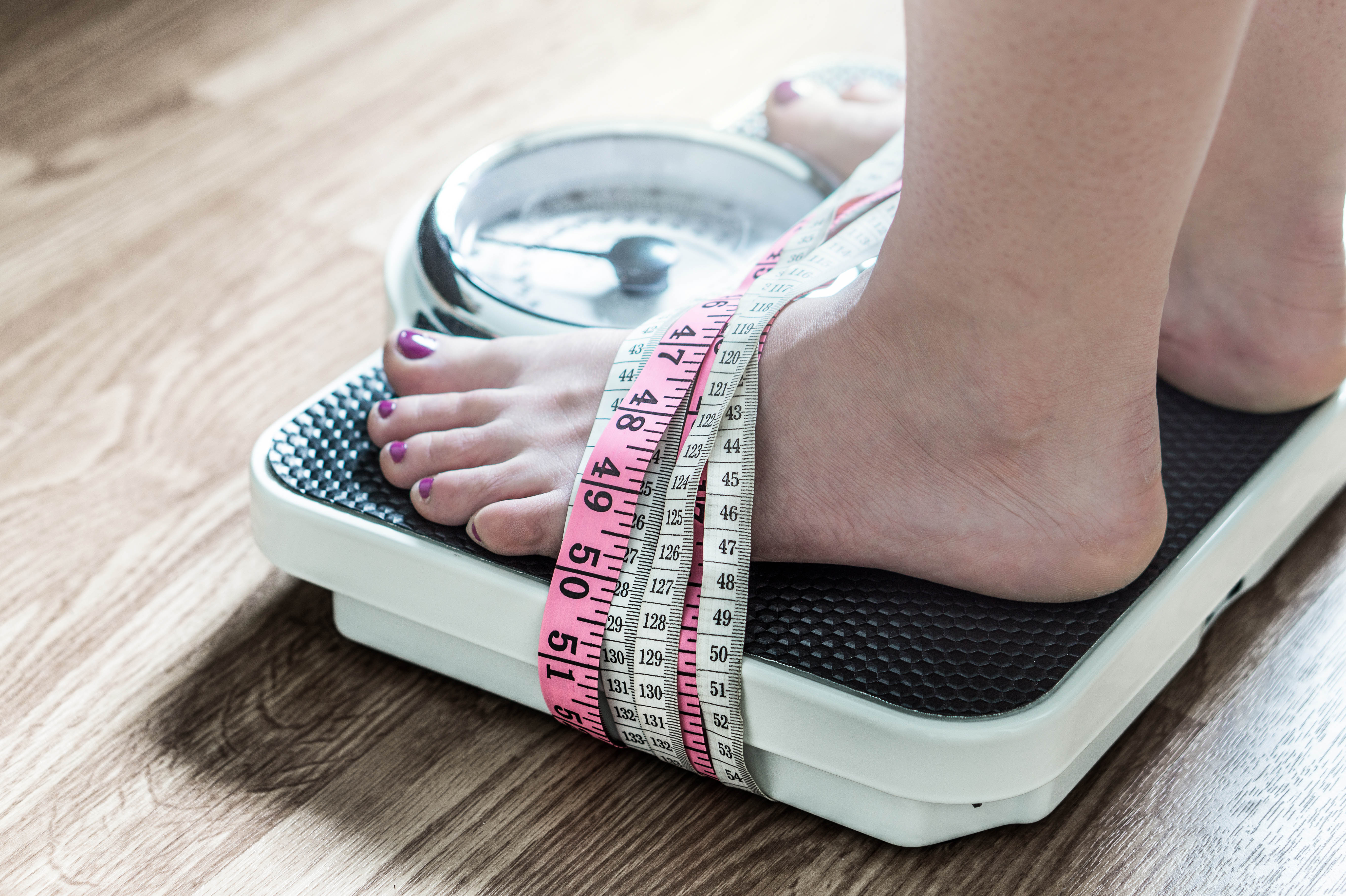 Close-up of woman's feet as she stands on a scale, with tape measures wrapped around her feet and the scale Close-up of woman's feet as she stands on a scale, with tape measures wrapped around her feet and the scale